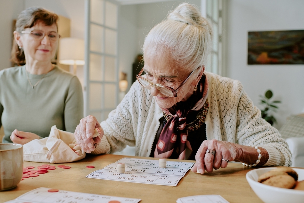 Senior Caucasian woman concentrating while playing bingo at table with middle aged Caucasian woman observing, both sitting indoors engaging in leisure activity together