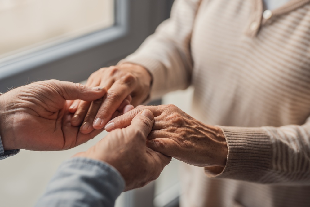 Elderly couple share a tender moment as one person gently holds the other’s hands, conveying compassion, trust and emotional support in a warm home setting with soft natural light.