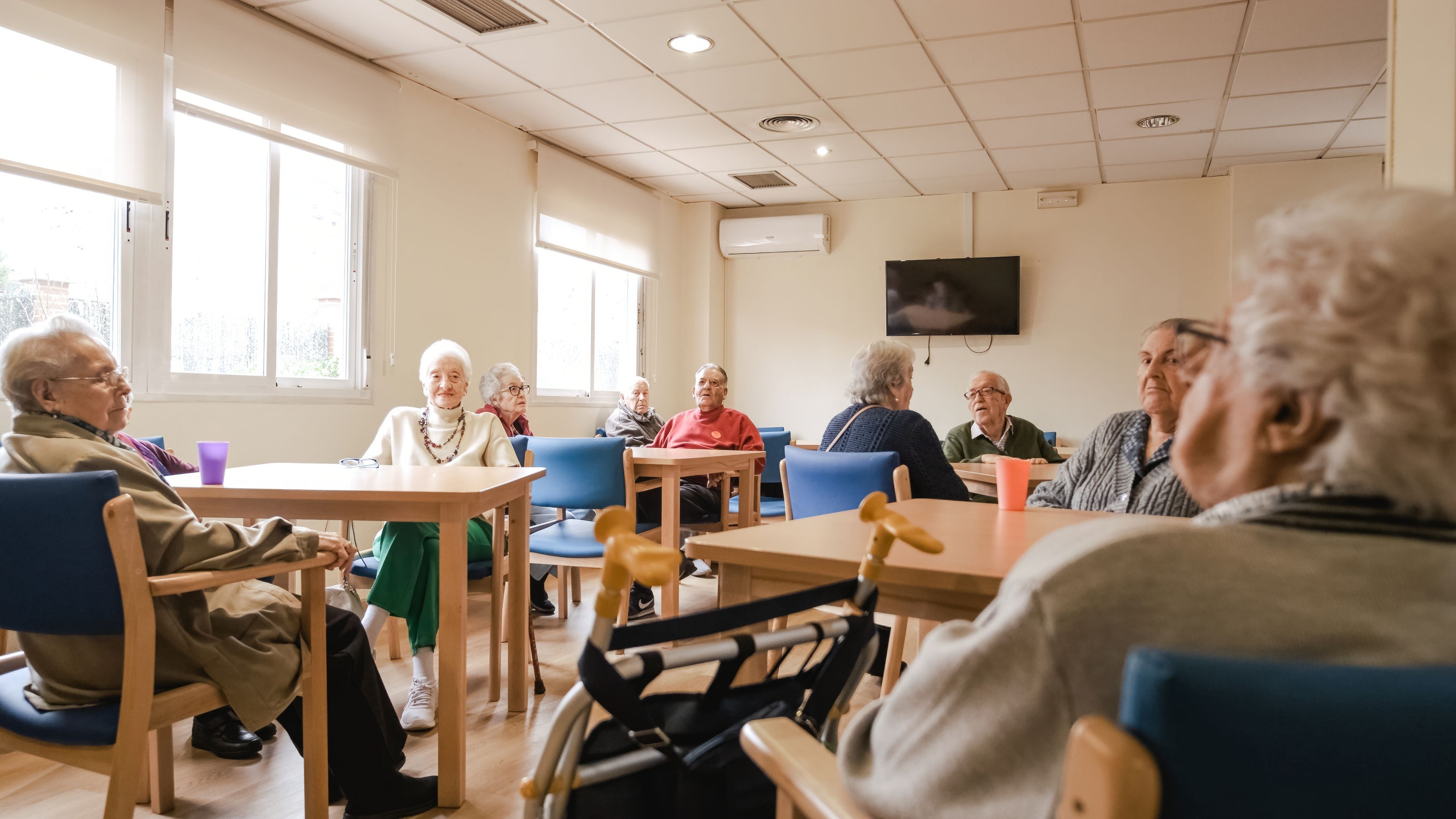 A room full of elderly couples smiling whilst sitting at tables in a large room with a TV on the wall