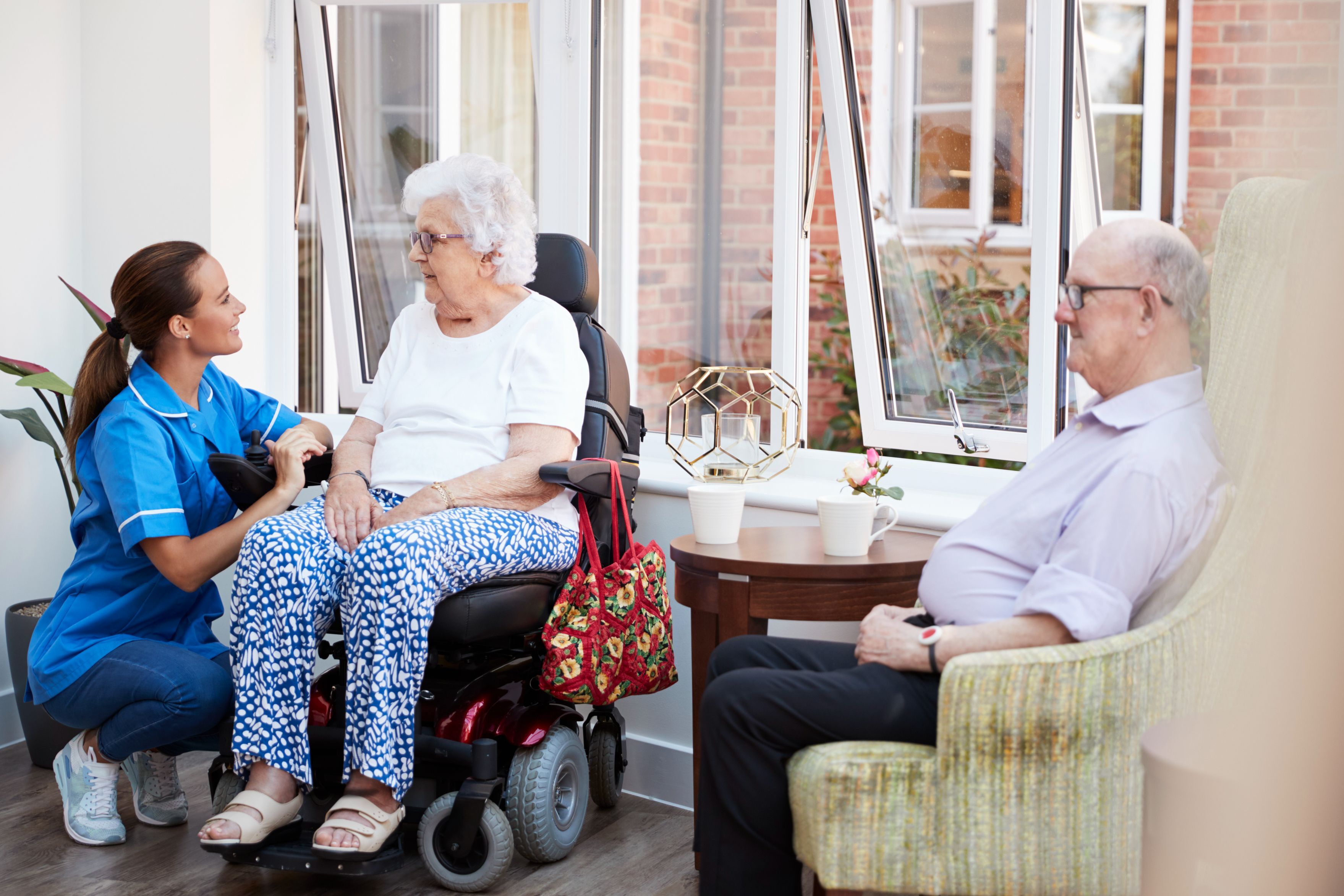 Women in wheelchair with elderly man in a chair next to her and younger care worker bending down to talk to the elderly lady.