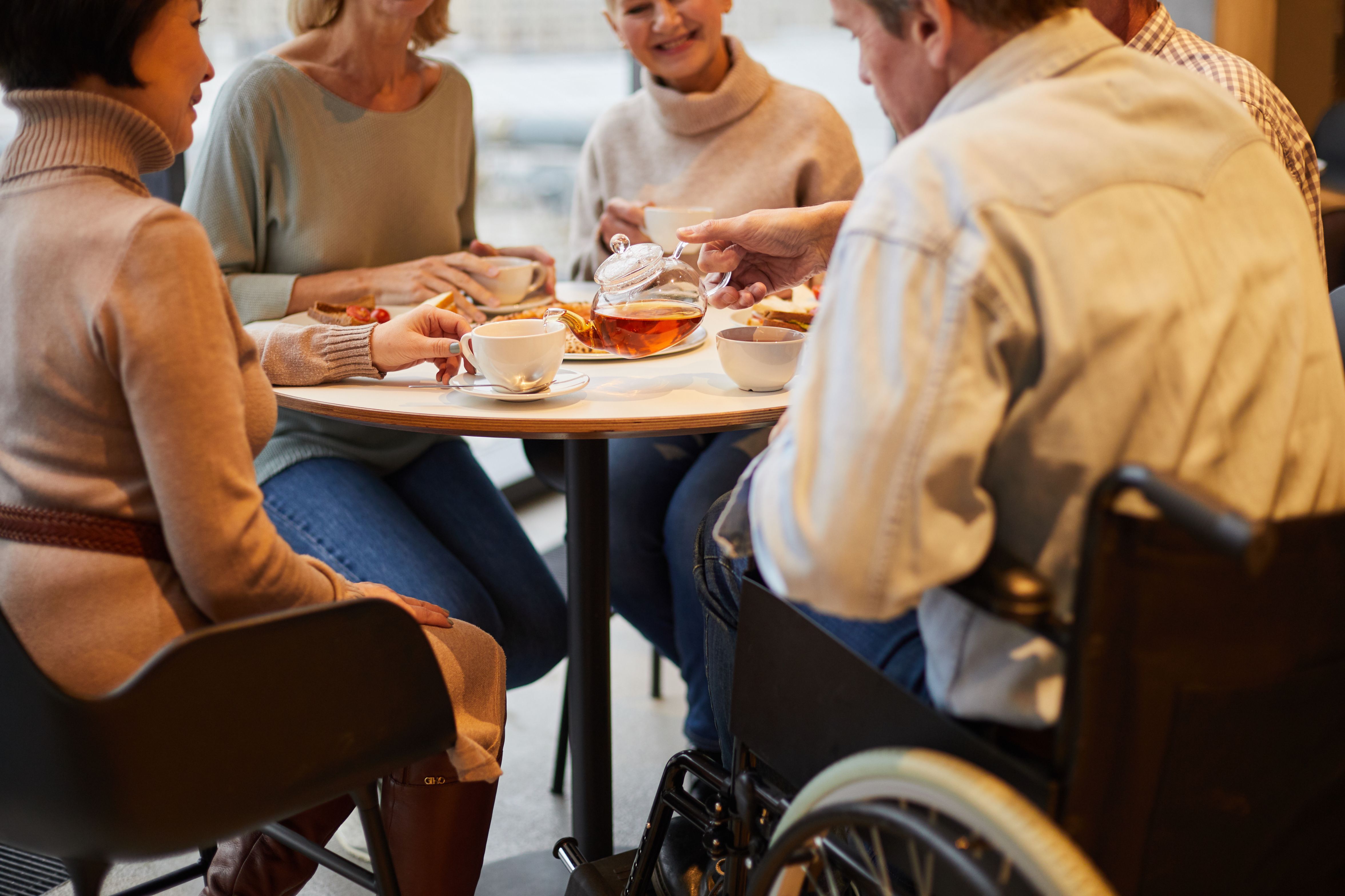 4 people sitting around a table enjoying tea. The man closest to the camera is in a wheelchair