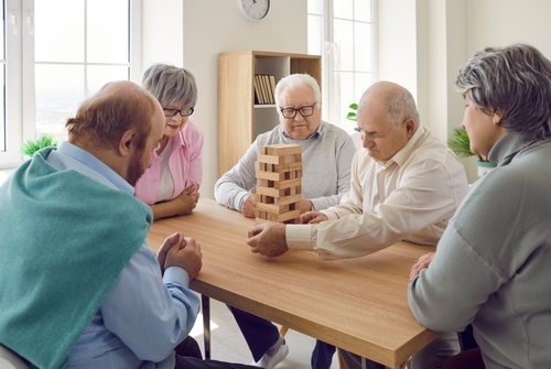 Group of senior men and women playing together with wooden building blocks for dementia therapy in nursing home enjoying activity sitting at the table. Leisure in retirement home concept.